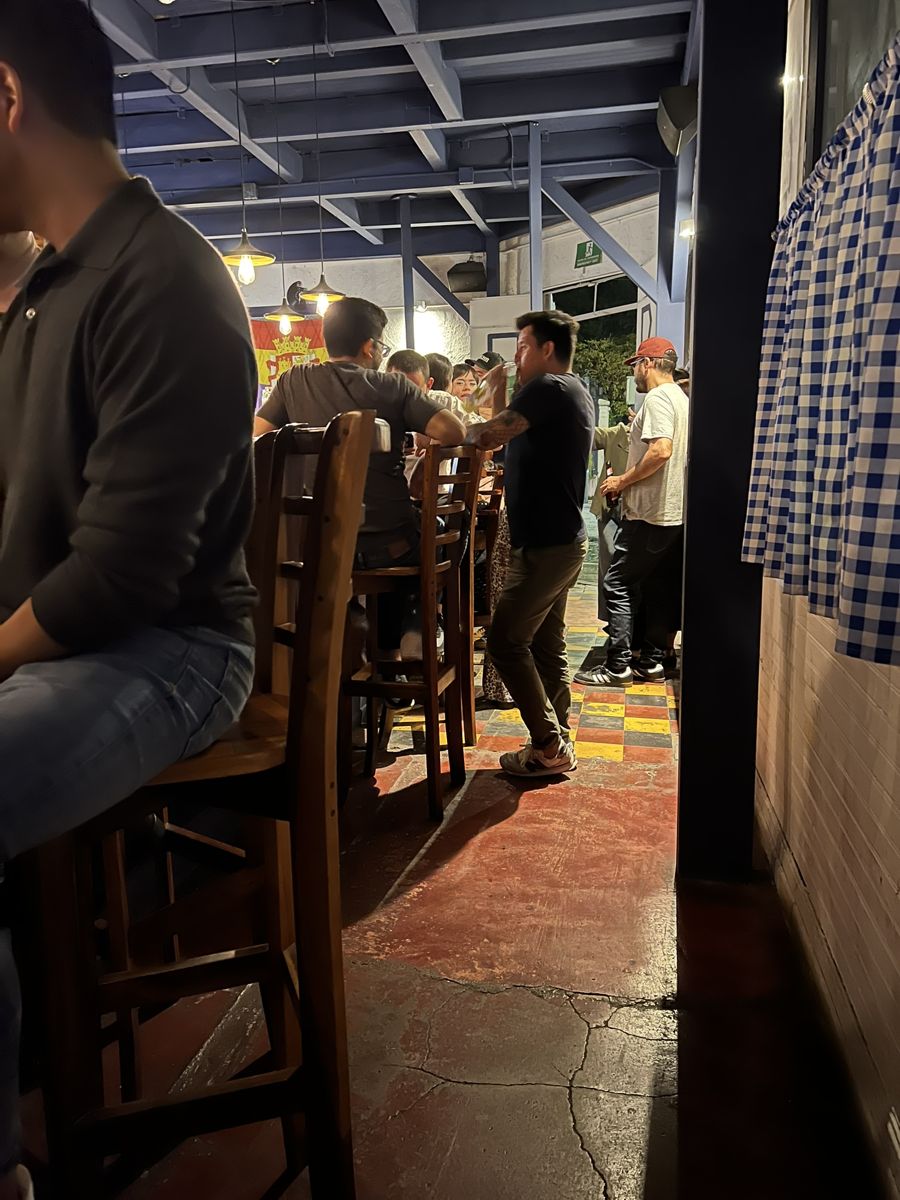 Pasillo del bar con bancos altos y cortinas a cuadros, lleno de gente
